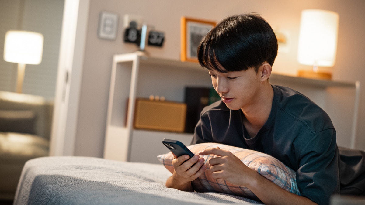 Um irmão jovem deitado na cama e olhando a tela de seu celular.