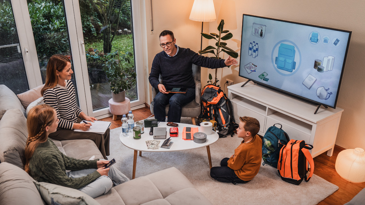 Um pai fazendo a adoração em família e ajudando a esposa e os dois filhos a se preparem para possíveis desastres. Eles estão assistindo ao vídeo “Como se Preparar para um Desastre?”. Eles têm na frente deles garrafas de água, dinheiro, uma Bíblia, enlatados, fita adesiva, mochilas de emergência e outros itens.