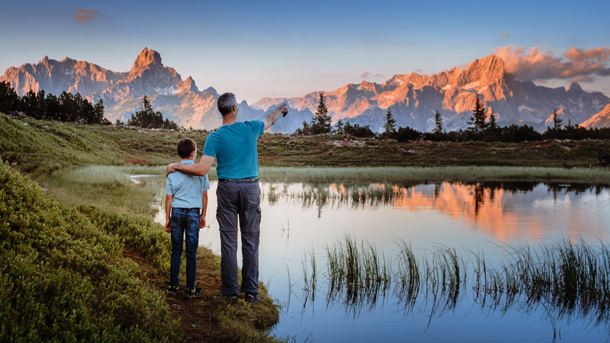 Um pai com seu filho jovem estão em pé diante de uma paisagem verde, olhando para uma cadeia de montanhas à distância.