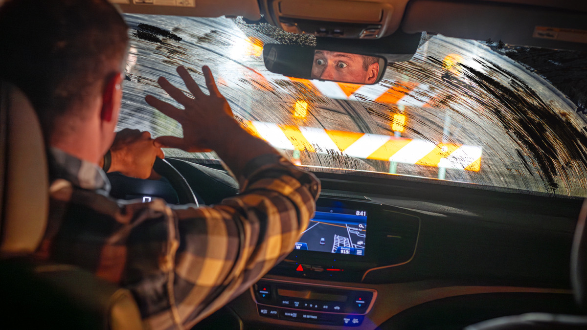 Homem dirigindo à noite com o para-brisa sujo e riscado, mal enxergando uma barreira à frente, o rosto assustado refletido no retrovisor
