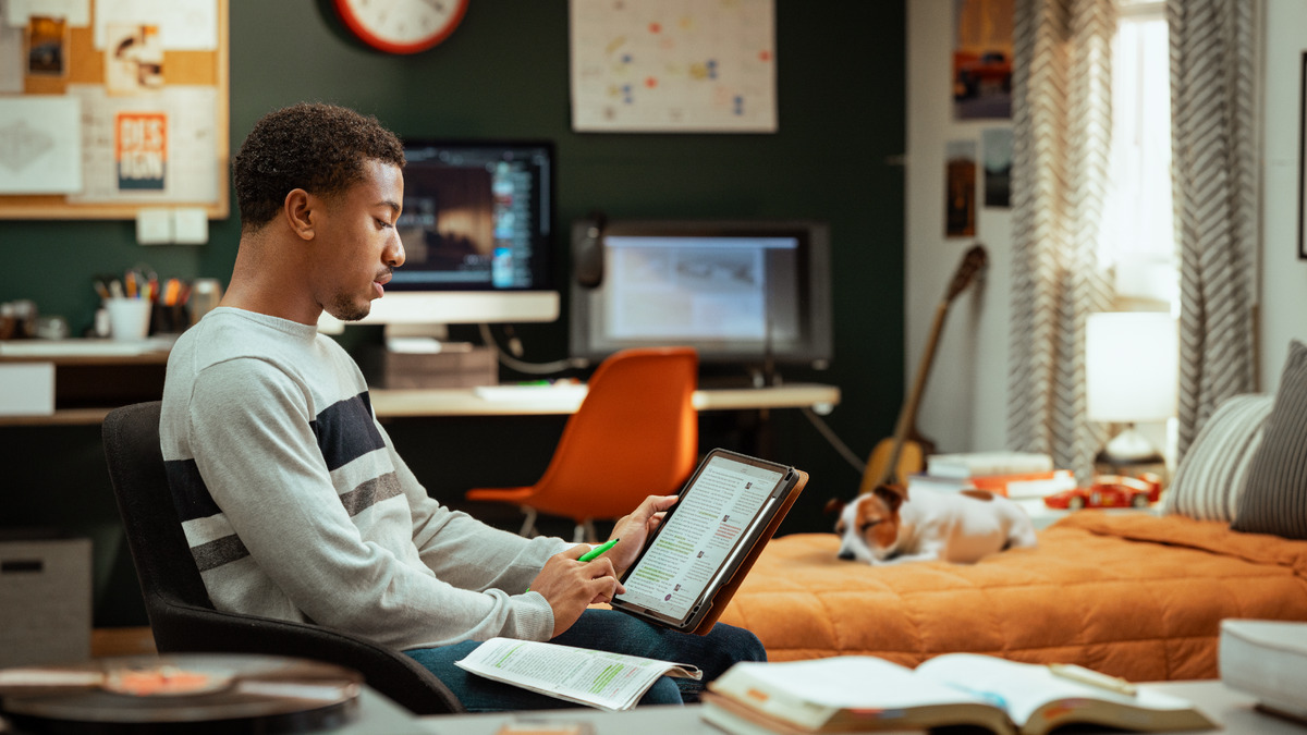 Um jovem estudante sentado no quarto prepara a reunião num tablet, com trechos destacados, uma Bíblia aberta na mesa e uma bancada de trabalho de design ao fundo