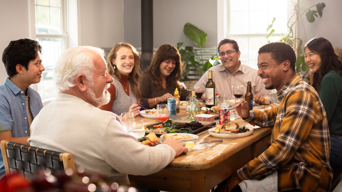 Sete irmãos de idades e origens diferentes riem juntos em volta de uma mesa de madeira, numa refeição em que há pratos variados e bebidas variadas, incluindo vinho, cerveja, refrigerante e água