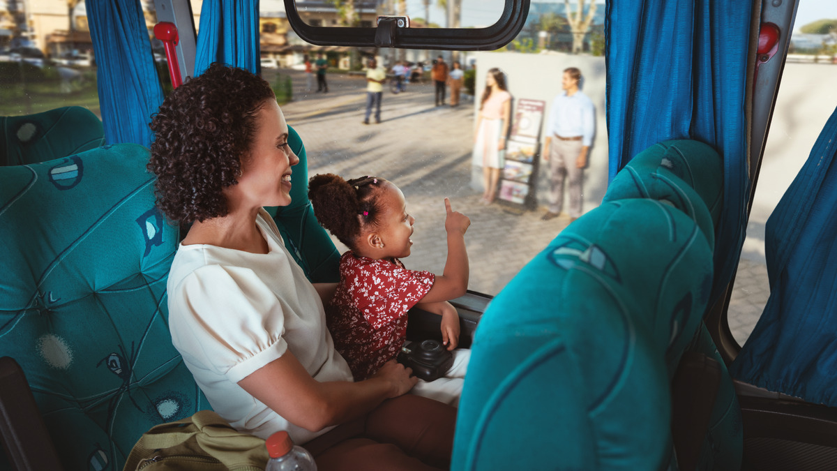 Dentro de um ônibus, uma mãe segura a filha pequena no colo e as duas sorriem ao ver pela janela um casal de Testemunhas de Jeová pregando com o carrinho na calçada