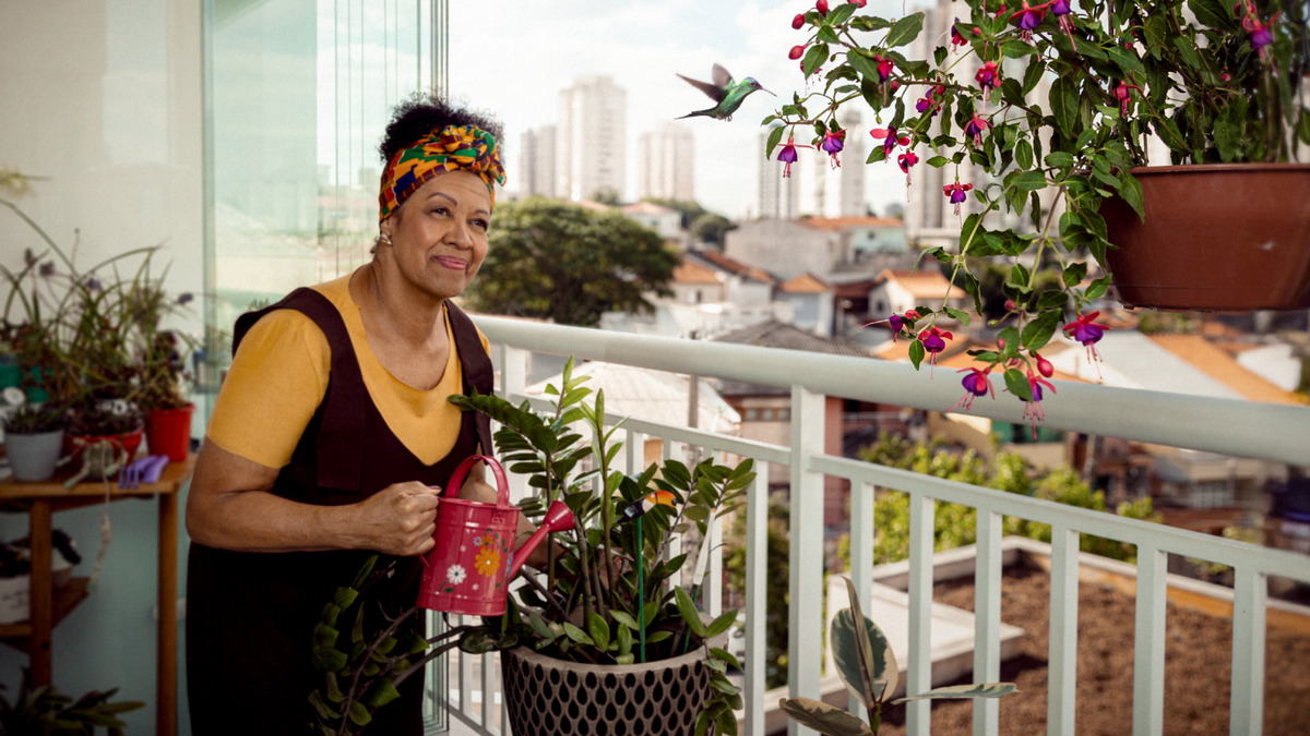 Uma irmã na varanda de sua casa, em uma cidade grande, regando as plantas e admirando um beija-flor que está se alimentando.